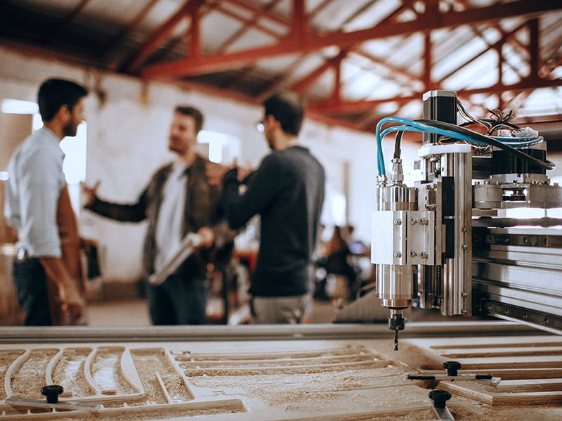 Maszyna CNC do obróbki w drewnie a w tle trójka rozmawiających mężczyzn