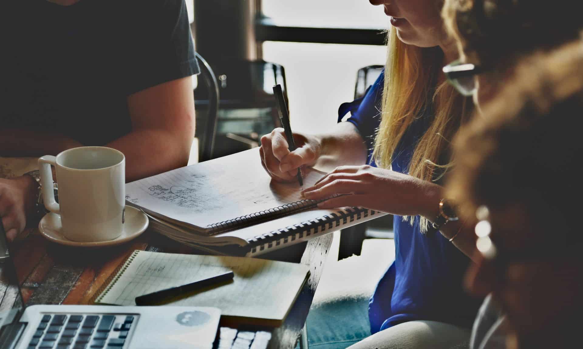 Kobieta robiąca notatki w notatniku obok którego leży kubek kawy i laptop