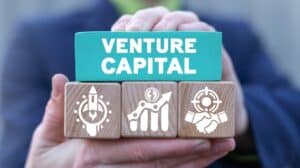 Osoba trzymająca drewniane klocki z napisem „Venture Capital” oraz ikonami symbolizującymi rozwój start-upów, wzrost inwestycji i partnerstwo biznesowe.