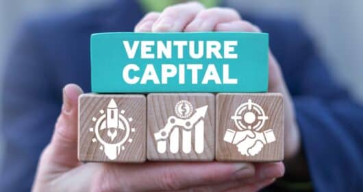 Osoba trzymająca drewniane klocki z napisem „Venture Capital” oraz ikonami symbolizującymi rozwój start-upów, wzrost inwestycji i partnerstwo biznesowe.