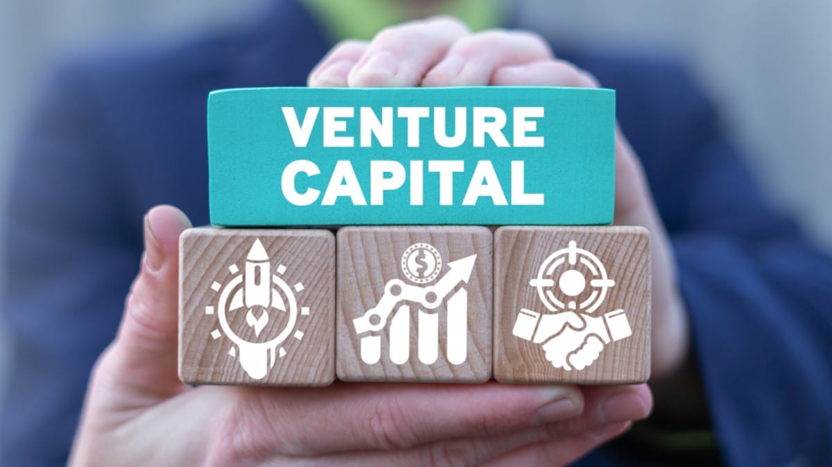 Osoba trzymająca drewniane klocki z napisem „Venture Capital” oraz ikonami symbolizującymi rozwój start-upów, wzrost inwestycji i partnerstwo biznesowe.