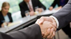 Zbliżenie na uścisk dłoni dwóch osób w garniturach podczas spotkania biznesowego; w tle widać rozmytych uczestników siedzących przy stole konferencyjnym