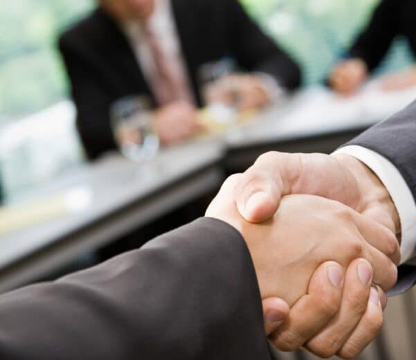 Zbliżenie na uścisk dłoni dwóch osób w garniturach podczas spotkania biznesowego; w tle widać rozmytych uczestników siedzących przy stole konferencyjnym