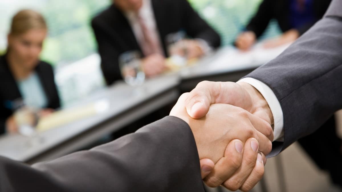 Zbliżenie na uścisk dłoni dwóch osób w garniturach podczas spotkania biznesowego; w tle widać rozmytych uczestników siedzących przy stole konferencyjnym
