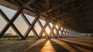 Zadaszony pasaż z metalową konstrukcją kratownicową, przez którą wpadają promienie zachodzącego słońca, rzucając długie cienie na posadzkę; w tle widoczne boisko sportowe i latarnie stadionowe.