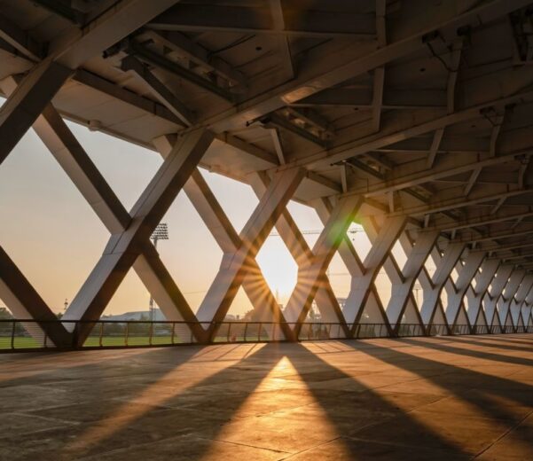 Zadaszony pasaż z metalową konstrukcją kratownicową, przez którą wpadają promienie zachodzącego słońca, rzucając długie cienie na posadzkę; w tle widoczne boisko sportowe i latarnie stadionowe.