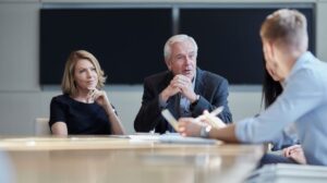 Cztery osoby siedzą przy stole konferencyjnym w nowoczesnej sali spotkań. Dwie osoby po jednej stronie stołu słuchają uważnie, podczas gdy osoba na pierwszym planie gestykuluje i mówi, trzymając notatki. Atmosfera przypomina formalne spotkanie biznesowe.