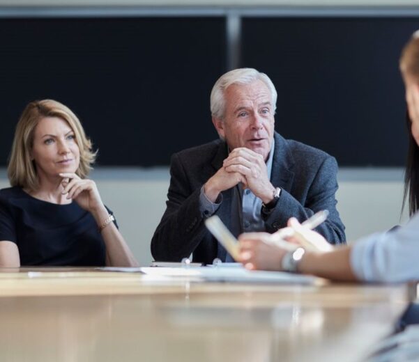 Cztery osoby siedzą przy stole konferencyjnym w nowoczesnej sali spotkań. Dwie osoby po jednej stronie stołu słuchają uważnie, podczas gdy osoba na pierwszym planie gestykuluje i mówi, trzymając notatki. Atmosfera przypomina formalne spotkanie biznesowe.